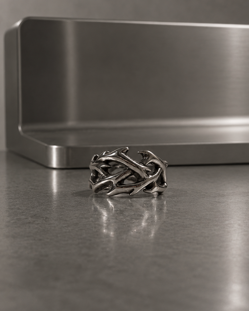 A silver-toned ring with an intricate, interwoven organic pattern, displayed on a reflective gray surface in front of a brushed metal rectangular block.
